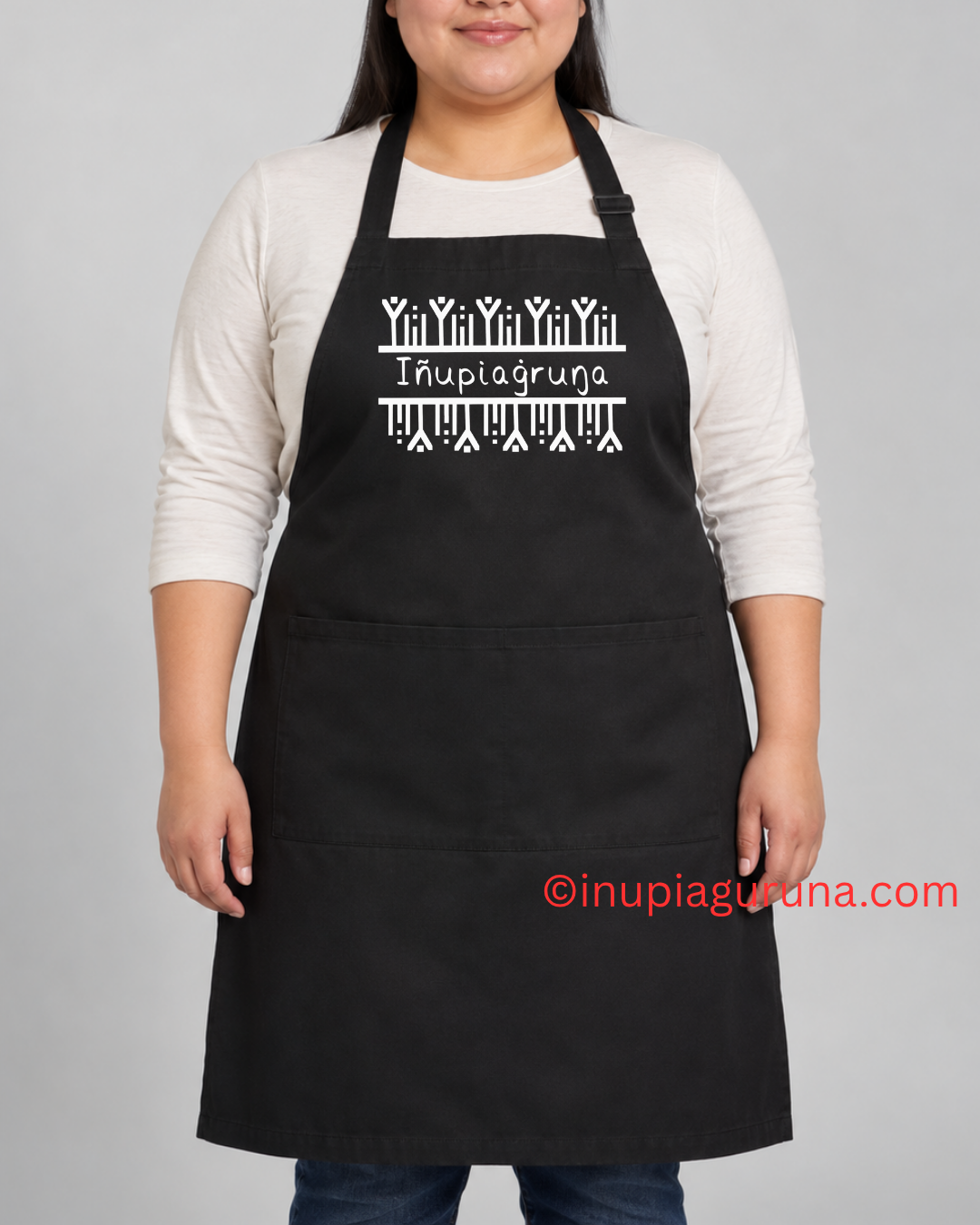 A person wearing a black apron over a white long-sleeve shirt, standing against a light gray background. The apron has a white Inupiagruna wordmark and decorative line art across the chest, with two front pockets.